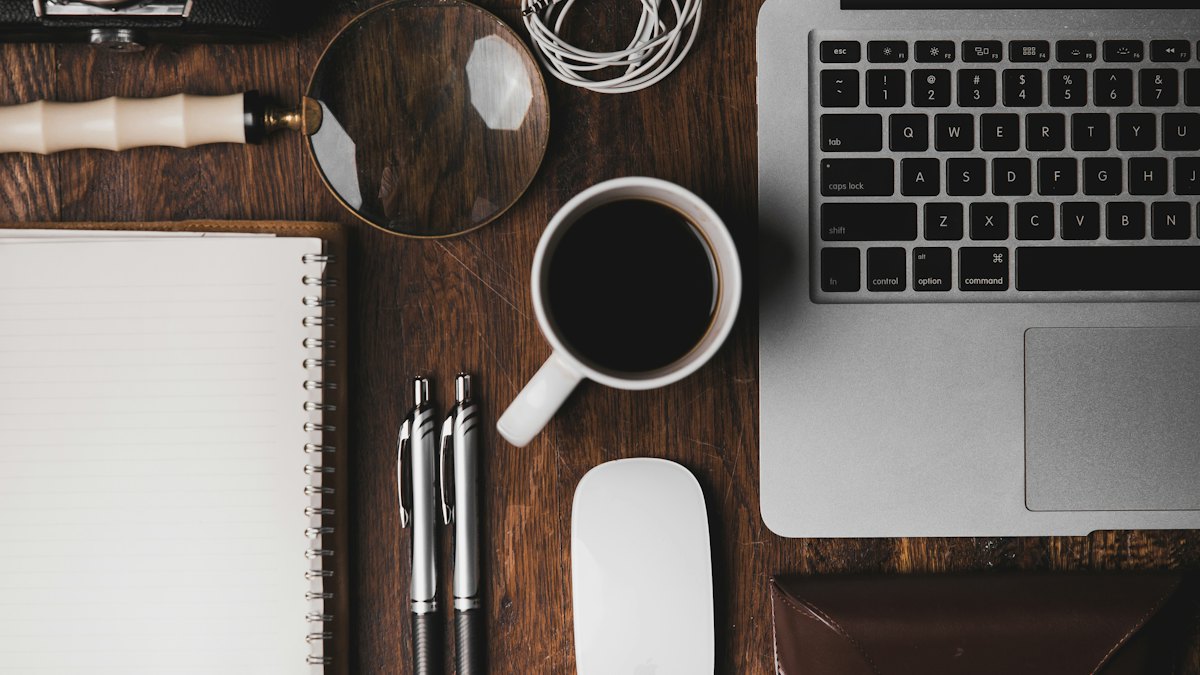 Flat lay of a desk with a laptop, notebook, coffee and pens